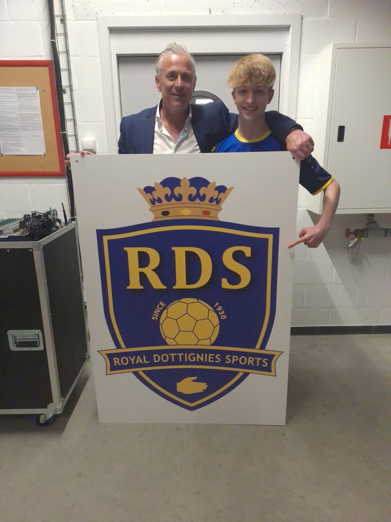 Two men pose behind a large sign bearing a blue-and-gold RDS crest with crown and the words 'ROYAL DOTTIGNIES SPORTS'. Behind-the-scenes area is visible with equipment cases nearby.