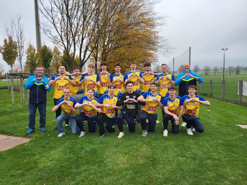 Youth soccer team posing on a grassy field in yellow and blue jerseys, making heart shapes with their hands; two coaches stand at the ends.