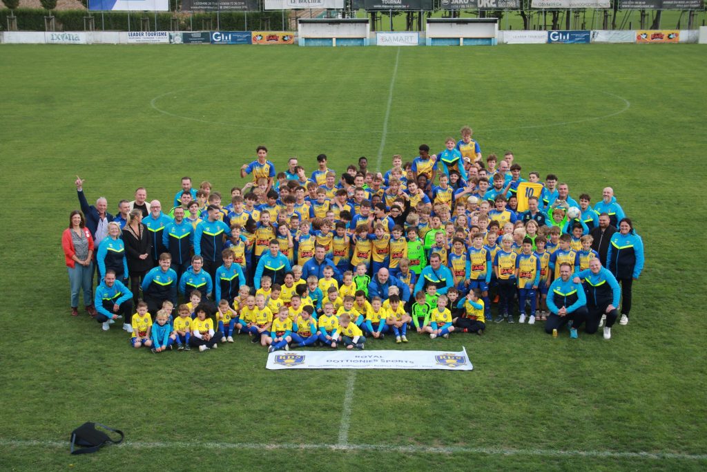 Group photo of a large youth soccer team in yellow and blue jerseys on a grass field with banners in the background.