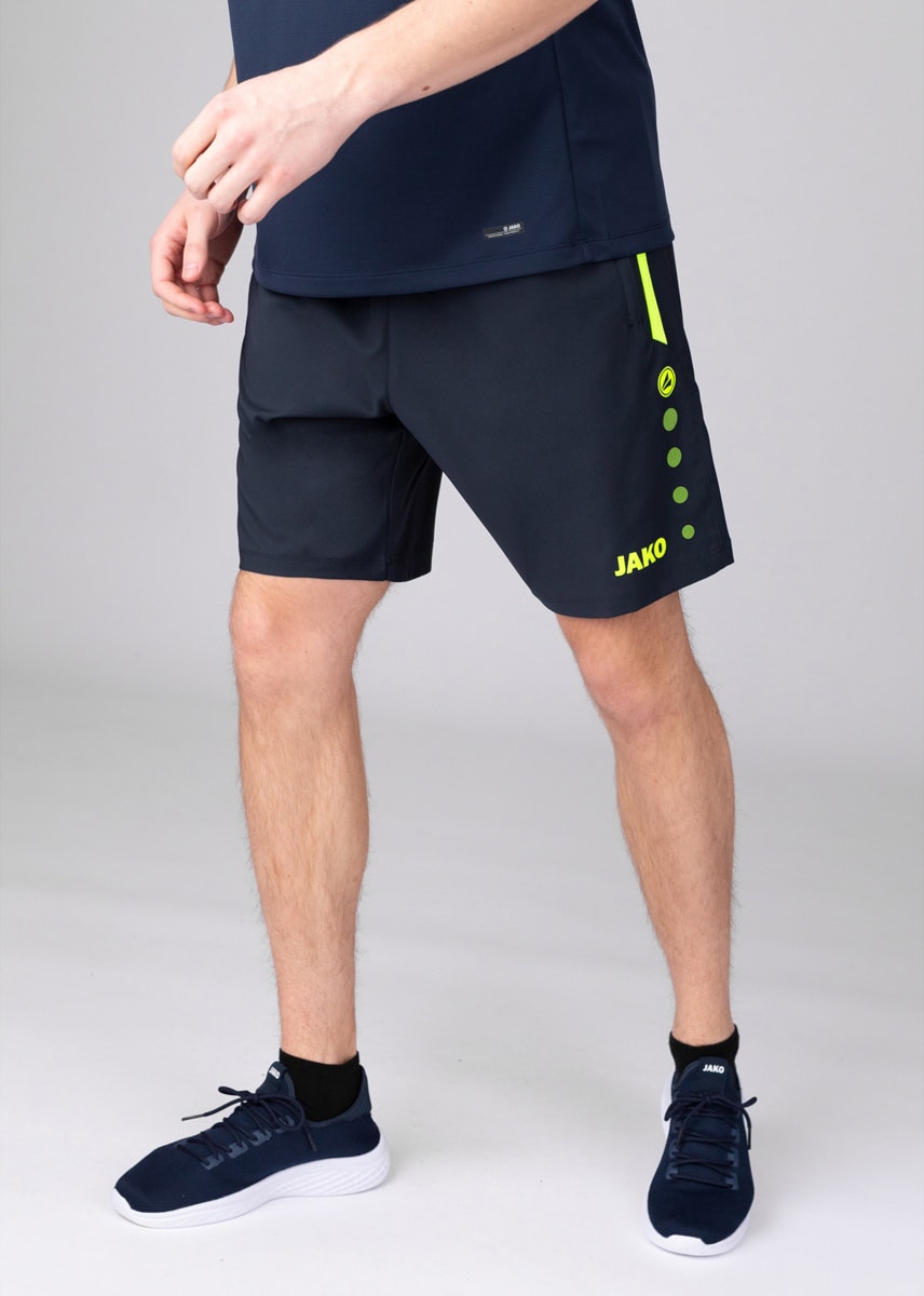 Man wearing navy athletic shorts with neon green accents and JAKO logo, standing in navy sneakers.