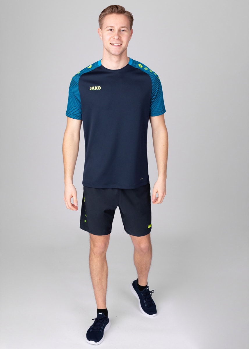 Young man in a navy athletic shirt with teal patterned sleeves and matching shorts, standing in a studio with a gray background and smiling.