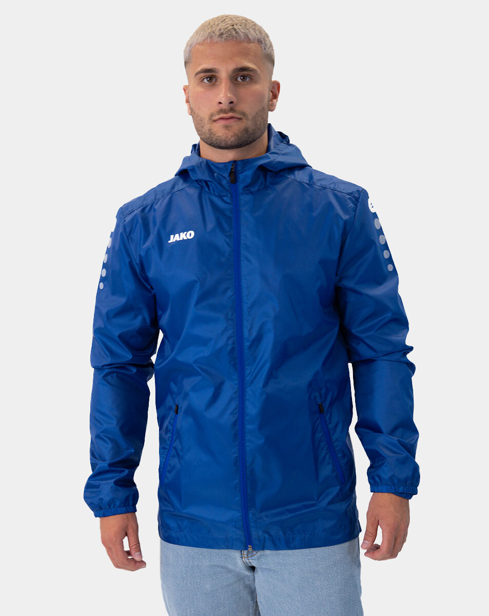 Man modeling a blue JAKO windbreaker with hood, front zipper, and pockets, against a light background.
